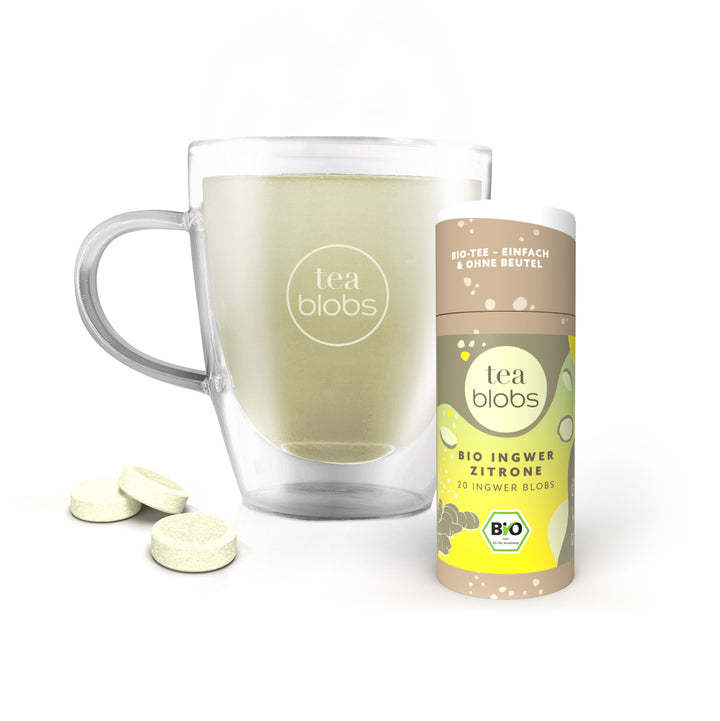 Glaskrug mit grünem Tee neben einer Packung Bio-Ingwer-Zitronen-Teeblöcke und drei einzelnen Teeblöcken.