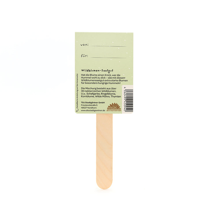 Holzstab mit einem grün bedruckten Etikett, das Wildblumensaatgut beschreibt, Felder für „von“ und „für“ enthält und Informationen über die enthaltenen Wildblumenarten gibt.