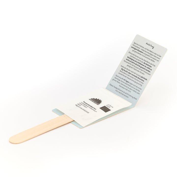 Ein Holzstab mit einer aufgeklappten Verpackung, in der sich eine Tüte mit Samen und eine gedruckte Anleitung in deutscher Sprache befinden.