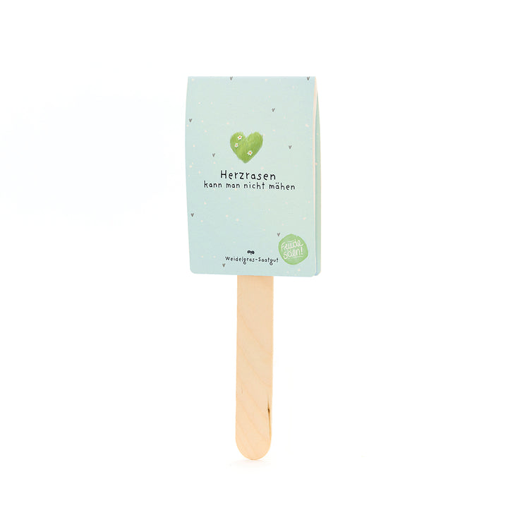 Holzstab mit hellblauem Schild, auf dem ein grünes Herz und der Text "Herzrasen kann man nicht mähen" sowie "Weidegras-Saatgut" steht.