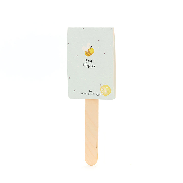 Holzstab mit hellblauem Etikett, darauf eine gezeichnete Biene und der Text "Bee Happy"; Wildblumen-Saatgut.