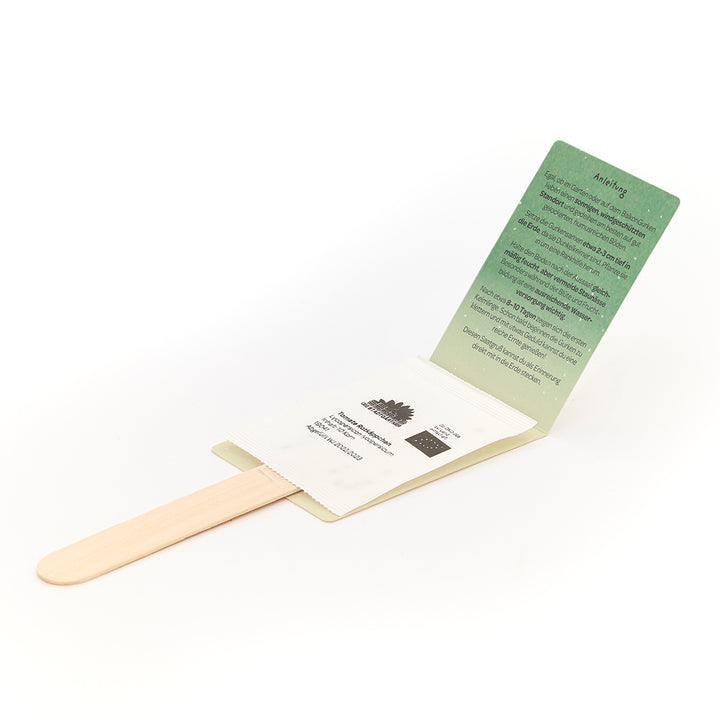 Holzstäbchen mit angebrachtem Umschlag, der eine grüne Anleitungskarte und ein weißes Saatgutpäckchen enthält.