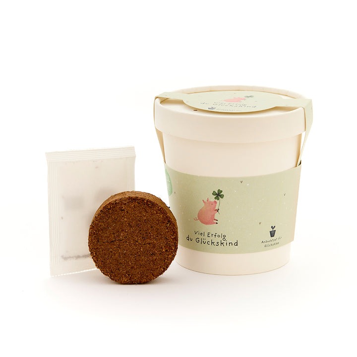 Pflanzset mit Becher, Erde und Samentütchen, auf dem Becher ein Schweinchen mit Kleeblatt und der Schriftzug "Viel Erfolg du Glückskind".