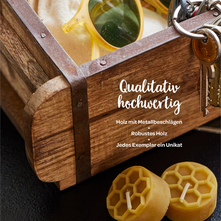 Holzkiste mit Metallbeschlägen, Sonnenbrille, Schlüsselbund und zwei Bienenwachskerzen; Text: "Qualitativ hochwertig – Holz mit Metallbeschlägen, Robustes Holz, Jedes Exemplar ein Unikat".