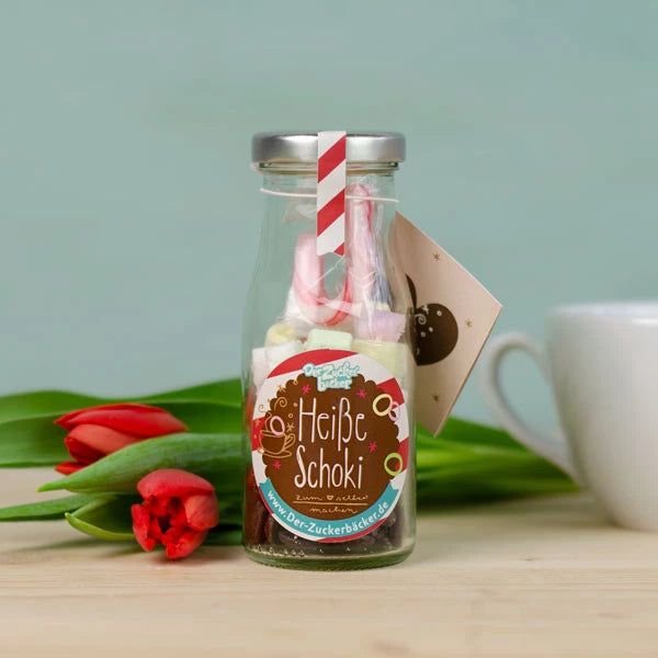 Flasche mit Etikett "Heiße Schoki" und Strohhalm, neben einer roten Tulpe und einer Tasse im Hintergrund.