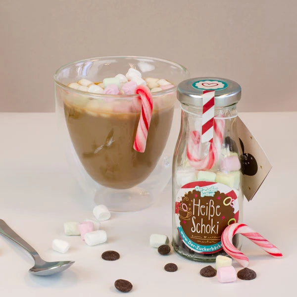 Ein Glas mit heißer Schokolade und Marshmallows, daneben eine Flasche mit der Aufschrift "Heiße Schoki" und Zuckerstangen, verstreute Marshmallows und Schokoladenchips im Vordergrund.