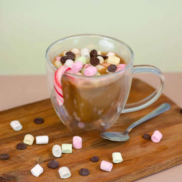 Heiße Schokolade in einer durchsichtigen Tasse mit Marshmallows, Schokoladenchips und Zuckerstange, serviert auf einem Holzbrett.
