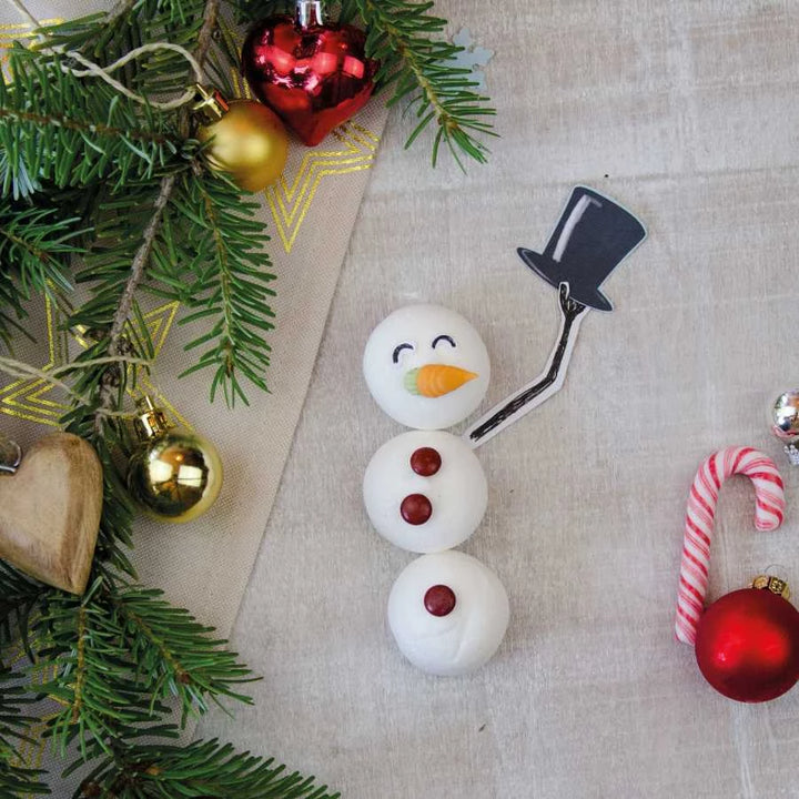 Ein Schneemann aus Marshmallows mit Zuckerhut, Schokolinsen und Karottennase liegt neben Weihnachtsdeko und Tannenzweigen auf einem Stoff.